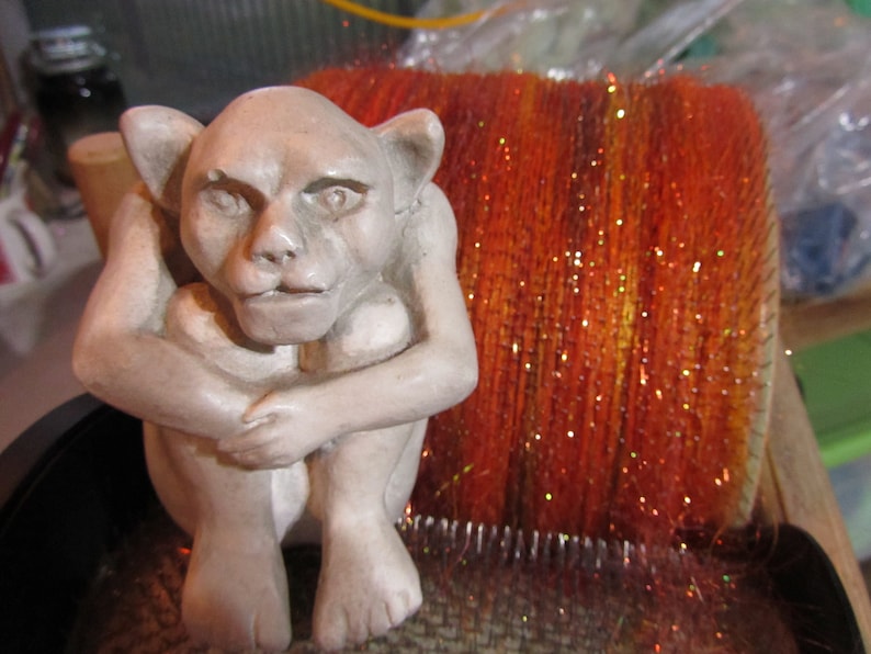 May include: A light gray, seated gargoyle figurine with pointed ears and a hunched posture, positioned in front of a large, cylindrical drum carder tool filled with vibrant, shimmering orange and red fiber with metallic gold flecks.