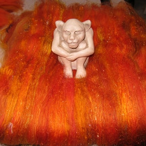 May include: A light beige, seated gargoyle figurine with pointed ears and a hunched posture, positioned atop a thick, textured layer of vibrant orange, red, and yellow wool roving fibers that feature subtle, shimmering metallic highlights.