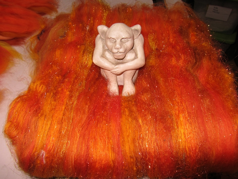 May include: A light beige, seated gargoyle figurine with pointed ears and a hunched posture, positioned atop a thick, textured layer of vibrant orange, red, and yellow wool roving fibers that feature subtle, shimmering metallic highlights.