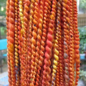 May include: A collection of multi-textured yarn strands in vibrant shades of orange, red, and yellow. The fibers feature various twists, including smooth, fuzzy, and metallic threads, with small embedded sequins reflecting light throughout the bundle.
