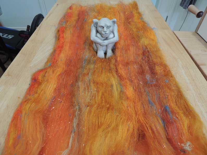 May include: A light gray, seated gargoyle figurine positioned atop a long, rectangular batt of blended wool fibers. The fiber batt features a vibrant color palette of orange, yellow, and red tones with subtle streaks of blue and white throughout.