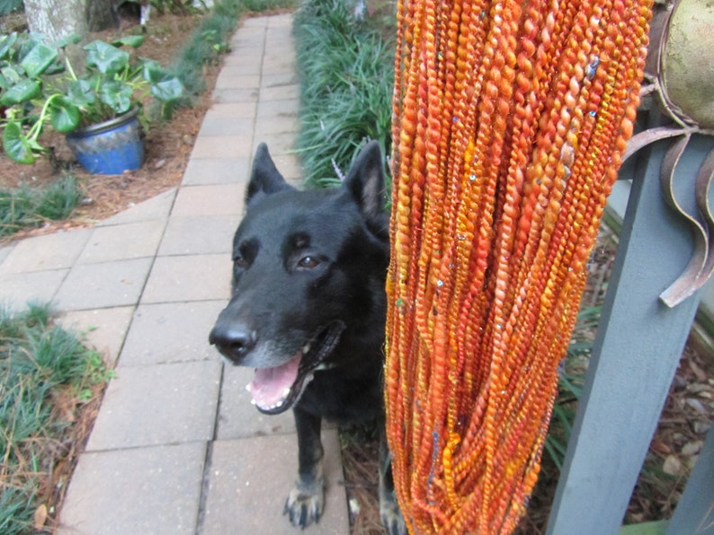 May include: A black dog with pointed ears and an open mouth stands next to a vertical bundle of twisted orange, yellow, and red yarn strands. The yarn is decorated with small, reflective sequins and beads woven throughout the textured fibers.