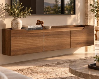May include: A wall-mounted wooden media console with three horizontal cabinet doors. The top surface holds a textured ceramic vase with greenery, a small bowl, a decorative knot object, and two books titled The Kinfolk Home and AT HOME.