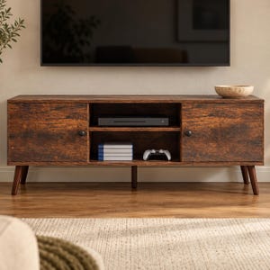 May include: A rectangular media console with a dark brown wood grain finish, featuring two side cabinets with small black knobs and a central open shelving unit containing a black electronic device, a stack of books, and a white game controller.