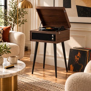 May include: A dark wood record player console with a hinged lid, a black speaker grille, and four tapered black legs. The turntable is visible inside the open top, featuring a black vinyl record with a yellow center label and a tone arm.