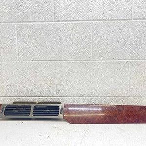 May include: A vehicle dashboard trim panel featuring a polished reddish-brown burl wood grain finish on the right side and a metallic silver section on the left containing two horizontal black air conditioning vents and integrated control buttons.