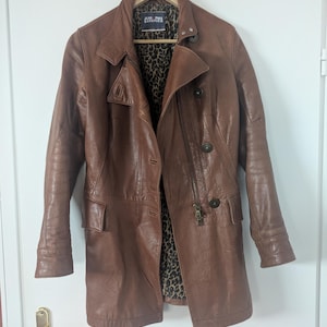 Puede incluir: Una chaqueta de cuero marrón con cuello de solapa, mangas largas y cierre de botones frontal. El forro interior presenta un estampado de leopardo en negro y marrón. Una etiqueta en el cuello dice Jean Paul Gaultier.