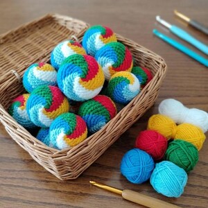 May include: A woven basket filled with small, crocheted spheres featuring a spiral pattern in red, yellow, green, blue, and white. Beside the basket are several solid-coloured yarn balls in white, yellow, red, green, and blue, plus crochet hooks.