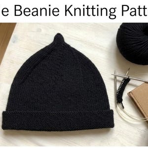 May include: A black knitted pixie beanie with a pointed top and a folded brim. The text Pixie Beanie Knitting Pattern is displayed above. Next to the beanie are two black yarn balls, a pair of circular knitting needles, and a small cardboard box.