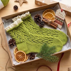 May include: A pair of lime green knit socks with a white ruffled cuff, presented in a cardboard gift box. The box contains dried orange slices, cinnamon sticks, pinecones, and a gift tag with the text Merry Christmas.