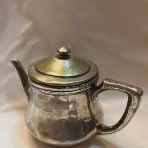 Op de afbeelding: Een kleine, doffe zilverkleurige theepot met een ronde vorm, een gebogen tuit en een stevig handvat. De theepot heeft een afneembaar deksel met een kleine knop in het midden. Het metalen oppervlak vertoont duidelijke tekenen van ouderdom en slijtage.