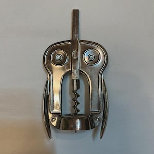 Könnte beinhalten: Ein silberfarbener Flügelkorkenzieher aus Metall mit einer zentralen Spirale, zwei seitlichen Hebeln und einer runden Basis. In der oberen Mitte des Geräts befindet sich ein kleines, rundes Logo mit dem Text "HIC", der deutlich eingeprägt ist.