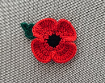 May include: A crocheted poppy flower applique featuring four vibrant red petals surrounding a solid black circular centre, accompanied by a single small dark green leaf extending from the upper left side of the flower head.