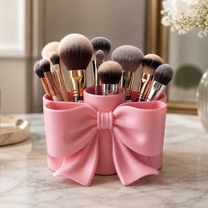 Op de afbeelding: Een lichtroze cilindrische make-up kwastenhouder met een opvallend gevormd strikontwerp aan de voorzijde. De houder bevat een verzameling diverse make-up kwasten met metalen hulzen en zachte, meerkleurige synthetische haren.