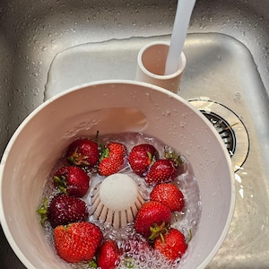Puede incluir: Un cuenco colador de bayas de plástico rosa claro que contiene fresas rojas frescas siendo enjuagadas bajo un chorro de agua. El cuenco cuenta con un cono de drenaje central elevado y un asa lateral, situado en un fregadero de acero.