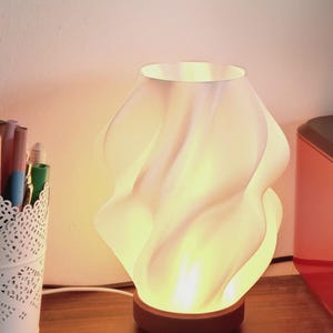 May include: A table lamp with a white, translucent, spiraling, wave-like shade that emits a warm, soft glow. The lamp features a circular, dark brown wooden base and is connected to a white power cord, providing illumination from within the structure.