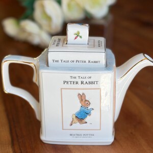 May include: A white ceramic teapot shaped like a book titled The Tale of Peter Rabbit. The lid features a small radish illustration. The front displays the book title and an illustration of Peter Rabbit in a blue jacket. Gold trim edges the handle.