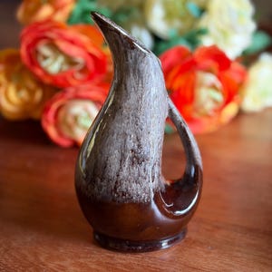 May include: A ceramic pitcher with a teardrop-shaped body and an integrated handle. The base is a deep, glossy brown, transitioning upwards into a textured, matte, light grey finish that extends to the pointed spout and the curved handle.
