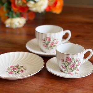 May include: Two white porcelain teacups and two matching saucers featuring a ribbed texture and a delicate pink rose floral pattern with green leaves. Each piece is finished with a thin gold trim along the scalloped edges of the rims.