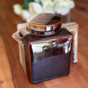 May include: A square ceramic teapot featuring a glossy dark brown finish on the main body and a textured, speckled beige and brown glaze on the lid, handle, and spout. The design includes a flat lid and a distinct angular handle and spout.