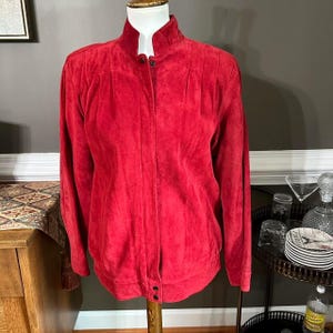 May include: A vibrant red suede jacket featuring a high collar, long sleeves, and a front button closure. The garment has vertical pleats extending from the shoulder area down the front, providing a structured fit with a soft, textured finish.