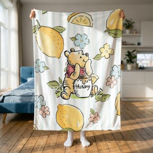 May include: A white throw blanket with light blue vertical stripes featuring a repeating pattern of yellow lemons, small blue and pink flowers, and a central illustration of Winnie the Pooh holding a honey pot with the word Hunny printed on it.