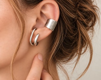 May include: A silver-toned ear cuff and a double-hoop earring set. The ear cuff is a wide, smooth band worn on the upper cartilage, while the double-hoop earring features two connected, rounded silver loops. The text ZioraJewelsCo is visible.