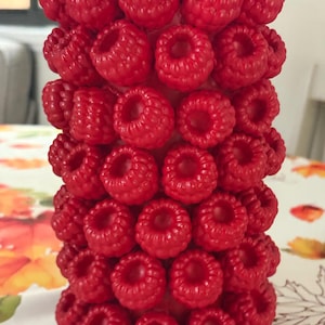 May include: A cylindrical object covered in a dense, repeating pattern of realistic, bright red raspberry-shaped embellishments. The surface is entirely textured with these small, rounded, berry-like forms, creating a uniform and tactile appearance.