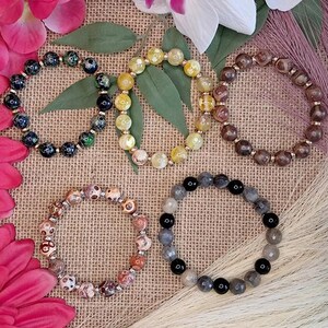 May include: Five beaded bracelets in various colors, including black, yellow, brown, and gray, are displayed. The bracelets are arranged on a textured surface, surrounded by pink and white flowers, and decorative elements. The bracelets appear to be made of natural stones.