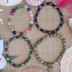 May include: Four beaded bracelets in various colors, including green, purple, and orange, are displayed on a textured, tan surface. The bracelets are adorned with round beads. Seashells and flowers surround the bracelets, adding a natural aesthetic.