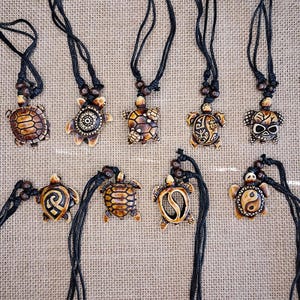 May include: A collection of handcrafted turtle pendant necklaces. Each pendant is a unique design, featuring intricate details and varying patterns in shades of brown and tan. The necklaces have black cords with wooden bead accents.