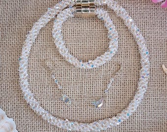 May include: A white beaded jewelry set, including a necklace, bracelet, and earrings. The necklace and bracelet have a gold magnetic clasp. The earrings feature a teardrop-shaped crystal pendant. The set is displayed on a textured, neutral-colored surface.