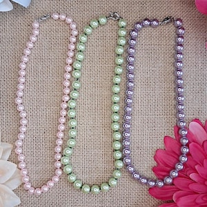 May include: Three beaded necklaces in shades of pink, green, and purple. Each necklace is composed of round beads and has a silver-colored clasp. The necklaces are displayed on a textured, neutral-colored surface, with floral accents.