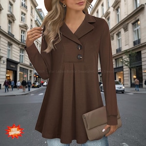 May include: A long-sleeve brown tunic top featuring a notch collar, two dark buttons on the front placket, and a pleated design that creates a flared silhouette. The garment has fitted sleeves with cuffs and a smooth, solid-colored fabric finish.