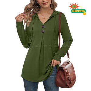 May include: An olive green long-sleeve pullover hoodie featuring a V-neckline with two dark buttons, a hood with drawstrings, and a pleated front detail. The garment is paired with blue denim jeans and a brown shoulder bag.