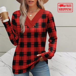 May include: A long-sleeve shirt featuring a red and black plaid pattern with a solid red V-neck collar. The neckline is accented with a vertical row of small gold-toned buttons. The shirt is paired with blue denim jeans and a white disposable cup.
