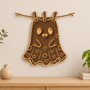May include: A wooden wall decoration featuring a dark brown ghost shape hanging from a horizontal line by three wooden clothespins. The ghost has a light wood border, two oval eyes, a small mouth, and a pattern of etched hearts and patches.
