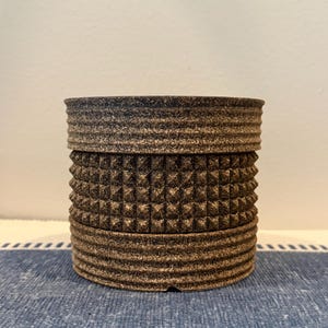 Op de afbeelding: Een cilindrische houder met een donkerbruine, gespikkelde afwerking. De buitenkant heeft drie horizontale banden: een gladde bovenrand, een middengedeelte met een patroon van piramidevormige noppen en een geribbelde onderkant.