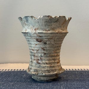 May include: A ceramic vase with a flared, uneven rim and a tapered body featuring horizontal ridges. The surface has a textured, matte finish in shades of off-white, gray, and brown, with visible rust-colored speckles and weathered detailing.