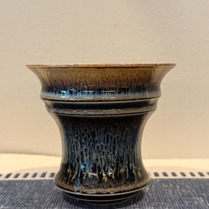 May include: A ceramic vase with a flared rim and a tapered body. The glaze features a gradient transition from a warm tan at the top to a deep, dark blue at the base, with white drip-like patterns running vertically throughout the surface.
