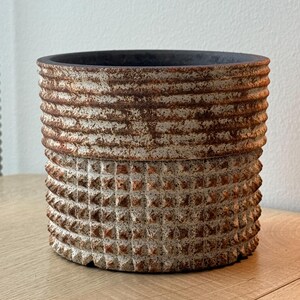 May include: A cylindrical planter featuring a two-part exterior design. The upper section has horizontal ridges, while the lower section displays a grid of raised square studs. The surface has a weathered, rust-coloured finish over a light base.