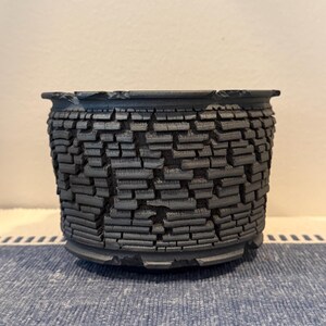 Puede incluir: Una maceta de cerámica gris carbón oscuro con un exterior texturizado que presenta un patrón repetitivo de pequeños segmentos rectangulares similares a ladrillos. El recipiente tiene una abertura ancha con un borde irregular.