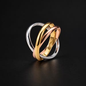 May include: A tri-color interlocking ring featuring three smooth, polished bands in silver, yellow gold, and rose gold tones. The three distinct metal bands are intertwined to form a single cohesive piece of jewelry with a reflective finish.