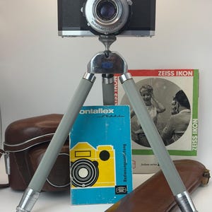 Könnte beinhalten: Eine silber-schwarze Zeiss Ikon Contaflex Kamera auf einem grauen Dreibeinstativ. Dazu gehören eine braune Ledertasche, ein braunes Lederetui, eine blau-gelbe Contaflex Super Bedienungsanleitung und eine Zeiss Ikon Broschüre.
