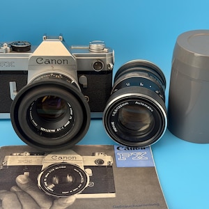 Pode incluir: Uma câmara de película Canon FX de 35 mm em prateado e preto com uma lente de 50 mm f/1.8, uma lente adicional Revue de 135 mm f/3.5 com estojo de plástico cinzento e um manual de instruções Canon FX com a imagem da câmara na capa.