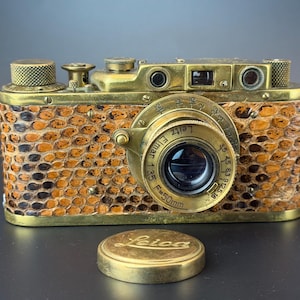 Peut inclure: Un appareil photo Leica vintage avec un corps en laiton et un revêtement texturé à motif peau de serpent marron et noir. L'objectif est un Leitz Elmar 1:3.5 F=50mm. Un bouchon d'objectif circulaire en laiton avec le logo Leica gravé.