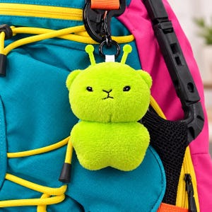 May include: A bright lime green plush keychain shaped like a small creature with two antennae, black embroidered eyes, and a simple mouth. The soft, fuzzy character is attached to a black plastic clip, which is fastened to a teal and pink backpack.