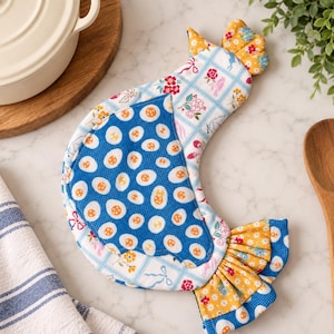 Könnte beinhalten: Ein Stofftopflappen in Hühnerform mit einem blauen Flügel mit weißem Eimuster, einem Körper mit hellblauem Gitter- und Blumenmuster sowie einem gelben Schwanz und Kamm mit kleinen Blumen- und Eimustern in Blau-, Weiß- und Gelbtönen.