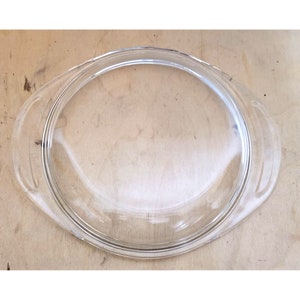 Puede incluir: Tapa de vidrio transparente con dos asas para una fuente para hornear. La tapa tiene forma ovalada y una superficie lisa y transparente.