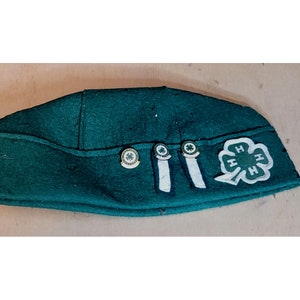 Puede incluir: Un sombrero de fieltro verde oscuro con un parche de trébol de cuatro hojas blanco y tres pequeños alfileres con tréboles de cuatro hojas verdes. El sombrero tiene una banda blanca con dos rayas blancas.